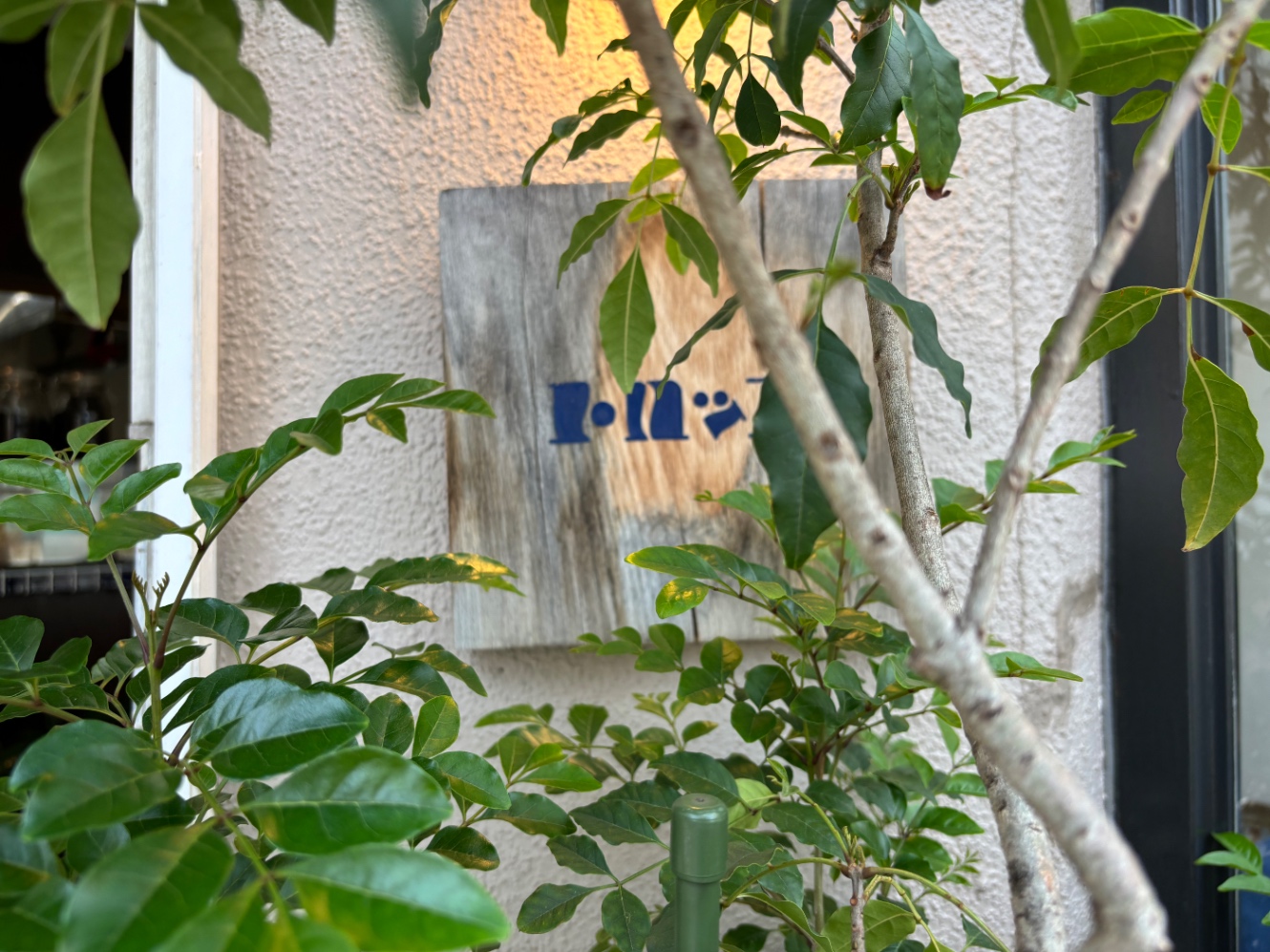 青字で木の看板にトロッコ、と書かれている　手前に植えてある植物の枝で一部看板が隠れている