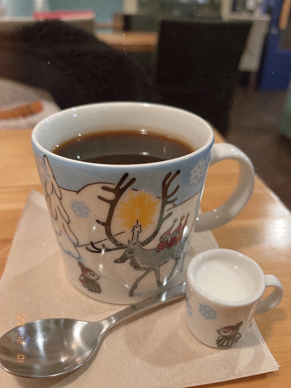 コーヒーが入ったマグカップの手前にスプーンと小さなミルクピッチャー。マグカップには冬のムーミン谷で頭にキャンドル、背中には3人の赤い人をのせたトナカイと思しき生き物とミイが向き合っている絵が描かれている。