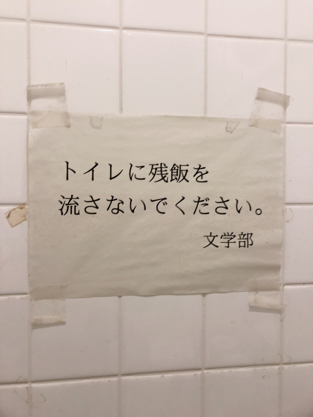 トイレの個室の壁の写真。白いタイルの壁に、「トイレに残飯を流さないでください　文学部」という貼り紙がされている。