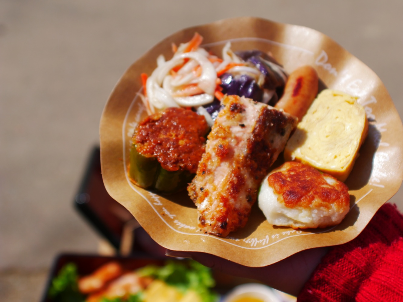 紙皿に盛られたおかず。南蛮漬け、ソーセージ、卵焼き、枝豆とチーズ入りの焼きはんぺん、サーモンのムニエル、ピーマンの肉詰め。