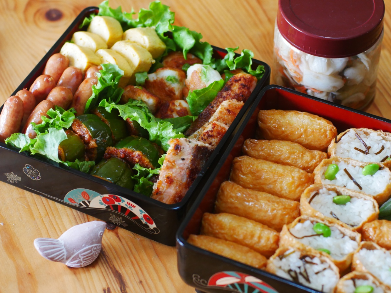 二段の重箱に詰められたお弁当。いなり寿司、卵焼き、ソーセージ、枝豆とチーズ入りの焼きはんぺん、ピーマンの肉詰め、サーモンのムニエルなど。重箱の横には瓶に入れられた揚げ茄子の南蛮漬けもある。