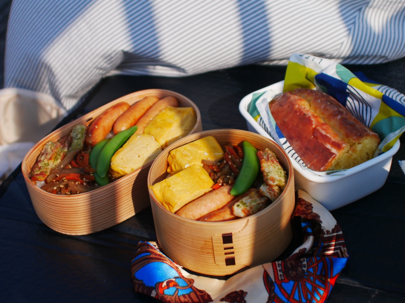 外でお弁当を広げている様子。白木の曲げわっぱがふたつ並び、ウインナー、卵焼き、ちくわの磯辺揚げ、きんぴら、スナップえんどうが詰められているのが見える。ホーローの小さな四角い容器にはウィークエンドシトロンが入っている。