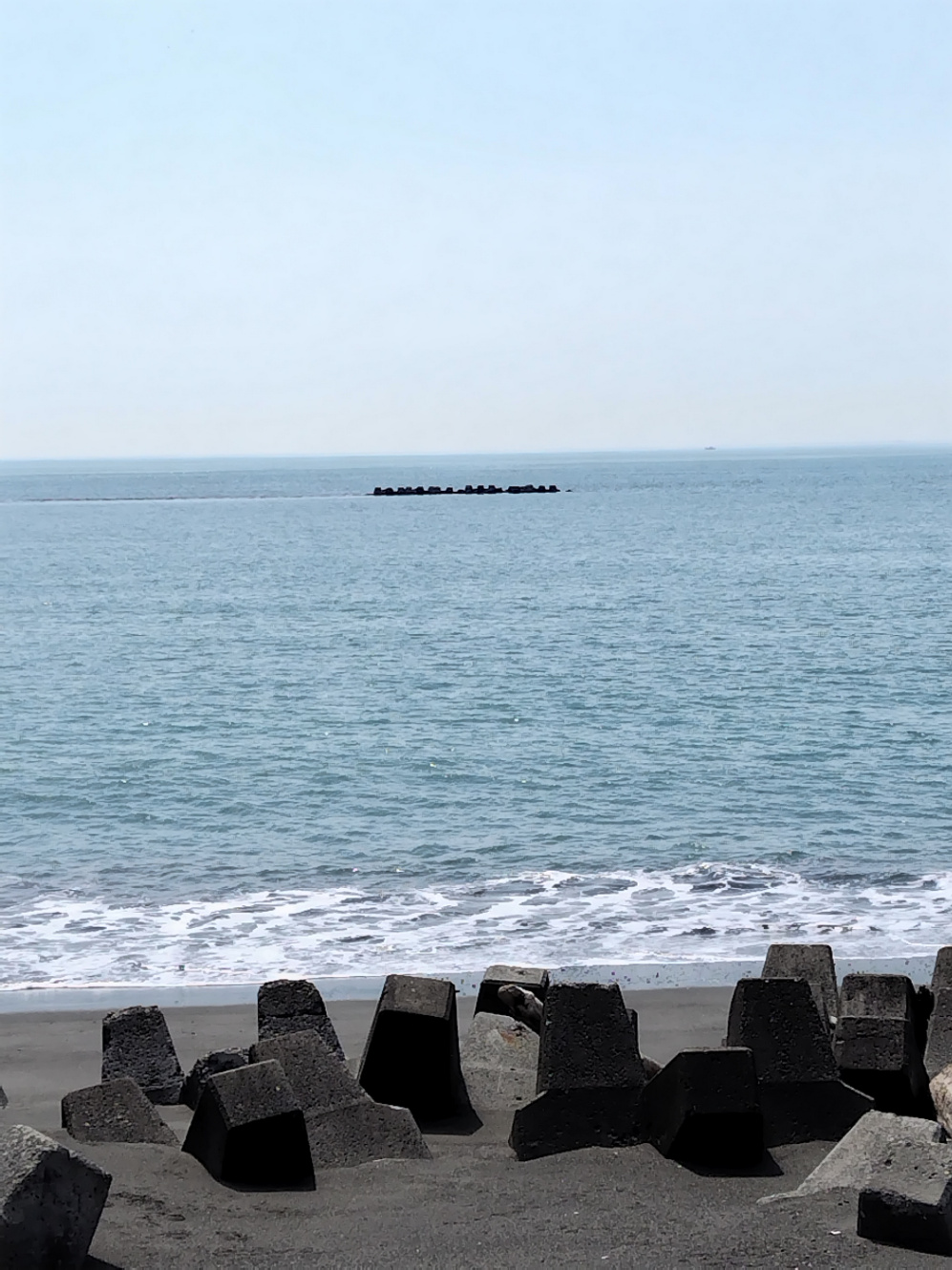 砂浜から海と波打ち際を撮影した写真。波は穏やかで、少し霞んだような青空に水平線がくっきりしている。