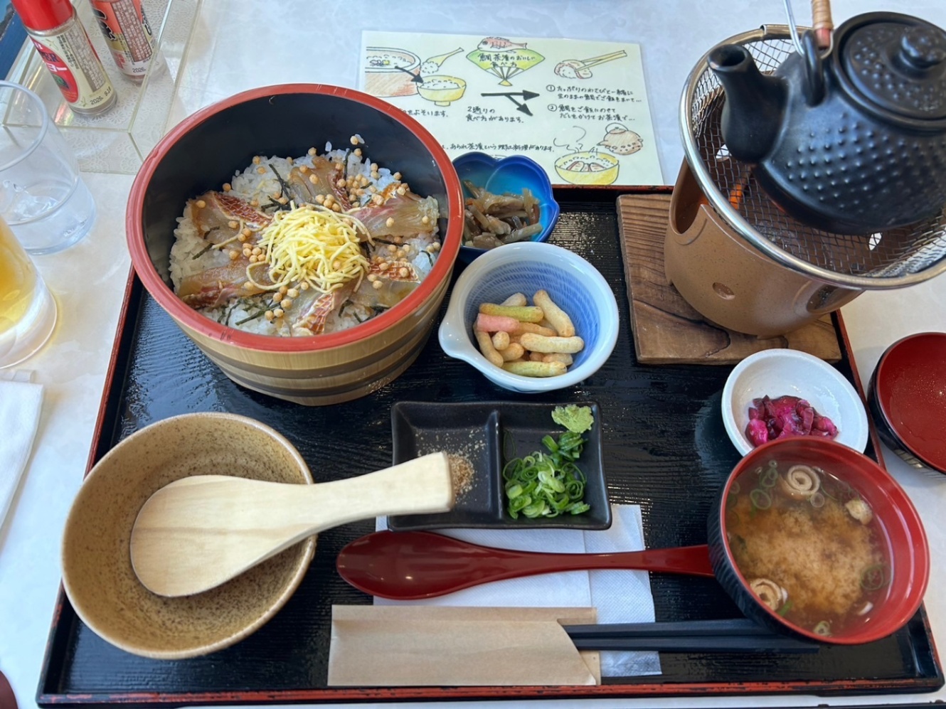 鯛めし。タイの切り身と錦糸卵ときざみのりがのったお櫃のご飯と、佃煮、お茶漬け用のあられ、おだし、漬物、味噌汁、薬味のネギとワサビとごま、茶碗としゃもじ、箸、スプーンが写っている