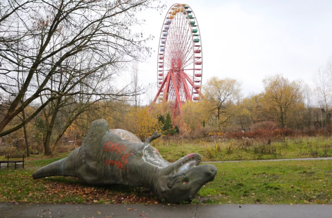 古びた遊園地の赤い観覧車。荒れた草むらに転がる、ペイントで落書きされたグレーの恐竜。