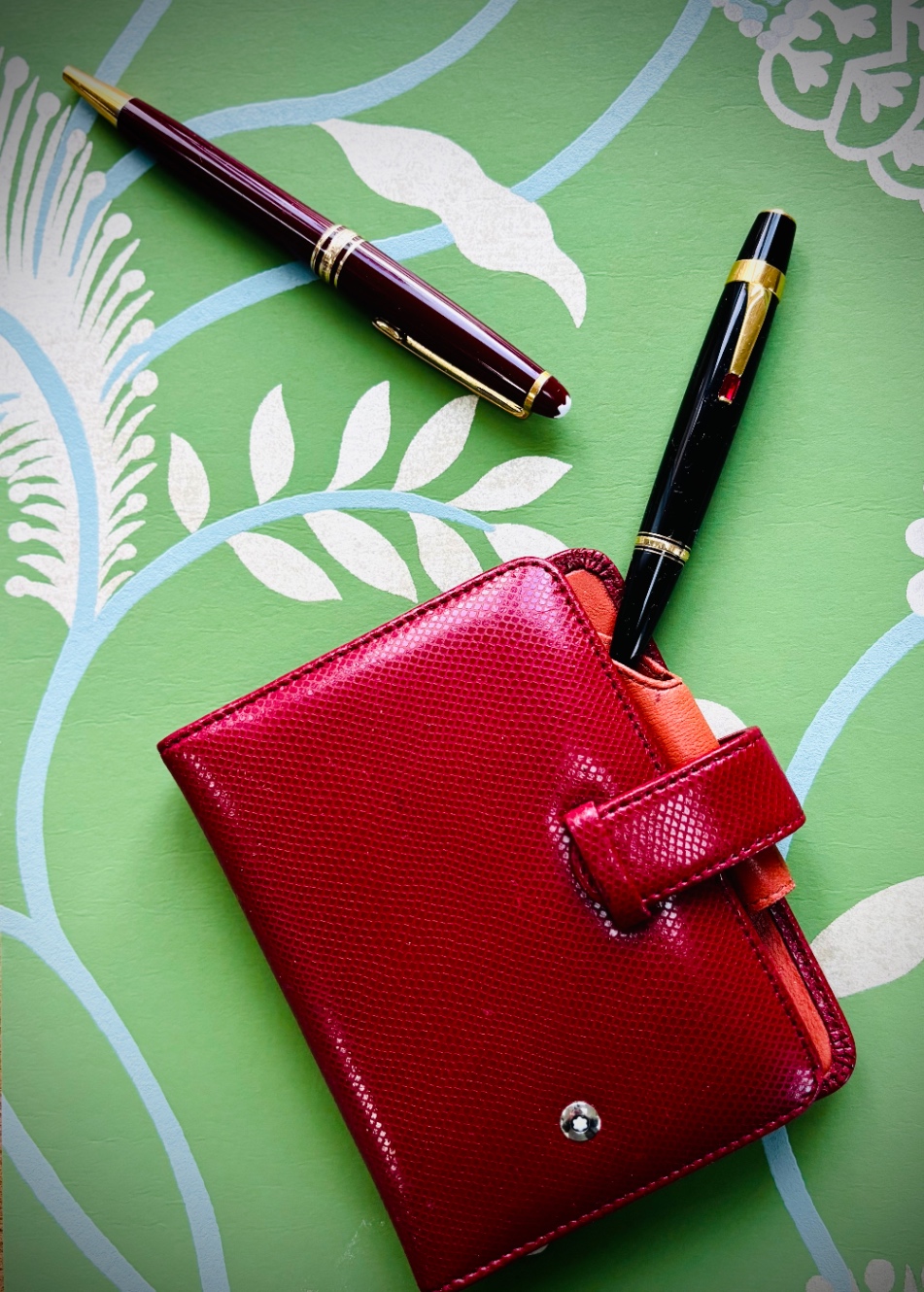 えんじ色の軸、ペン先とキャップの端、クリップ部分がゴールドのモンブランのボールペン。その横にはクリップ部分にルビー色の小さなジェムが付いた黒いモンブランのボールペン。そして真っ赤なグロッシーなレザー製のアドレスブック。こちらもモンブラン。