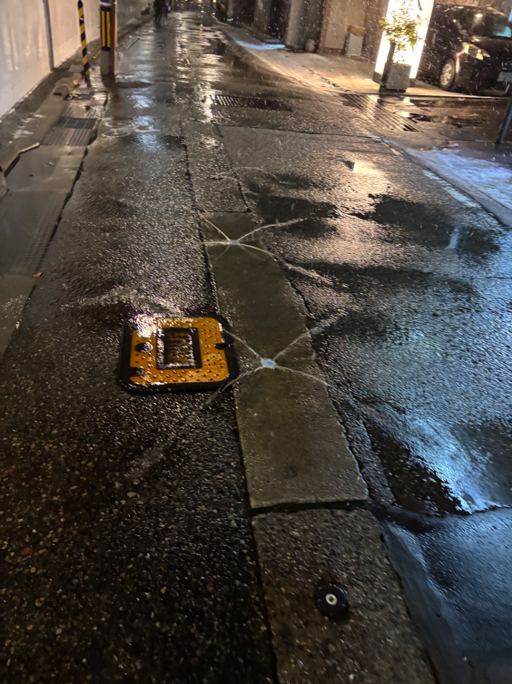 夜の金沢。濡れた路地裏。脇には雪が積もっているが、道路には1メートル間隔で設置された消雪パイプの穴から地下水が四方に噴射しており、まったく雪が見られない。