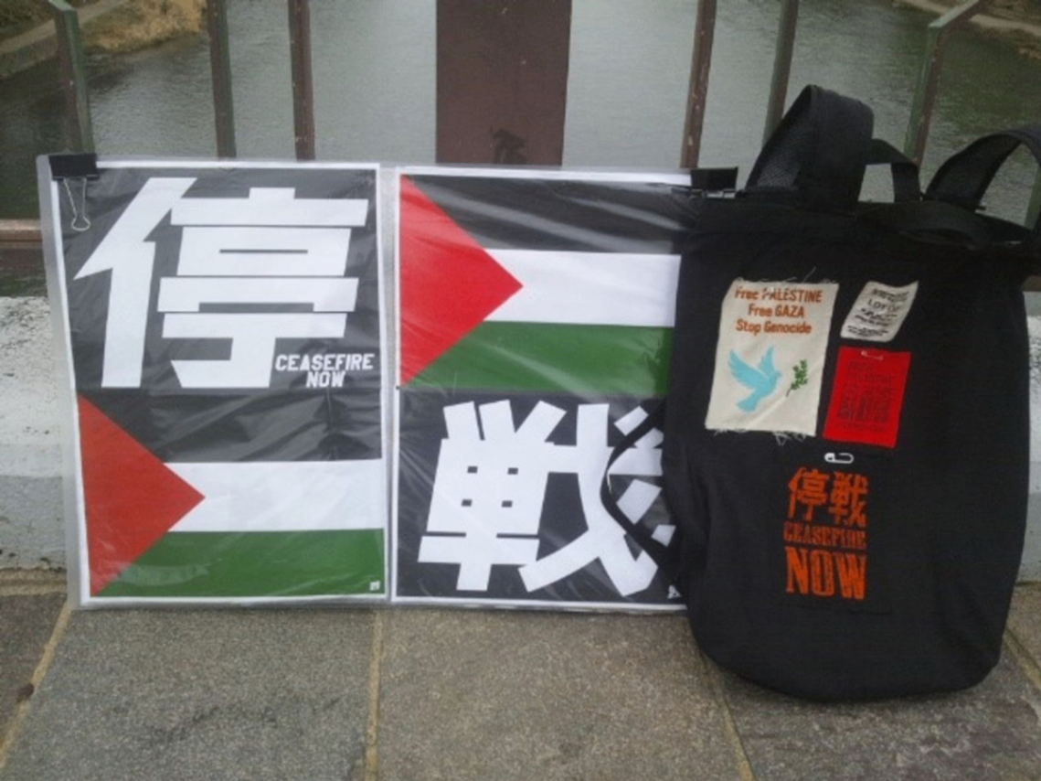 「停戦」の文字とパレスチナ国旗を収めたプラカード。右側に「停戦」や「Free Palestine」「Free GAZA」とプリントされたパッチを貼った黒いリュック。