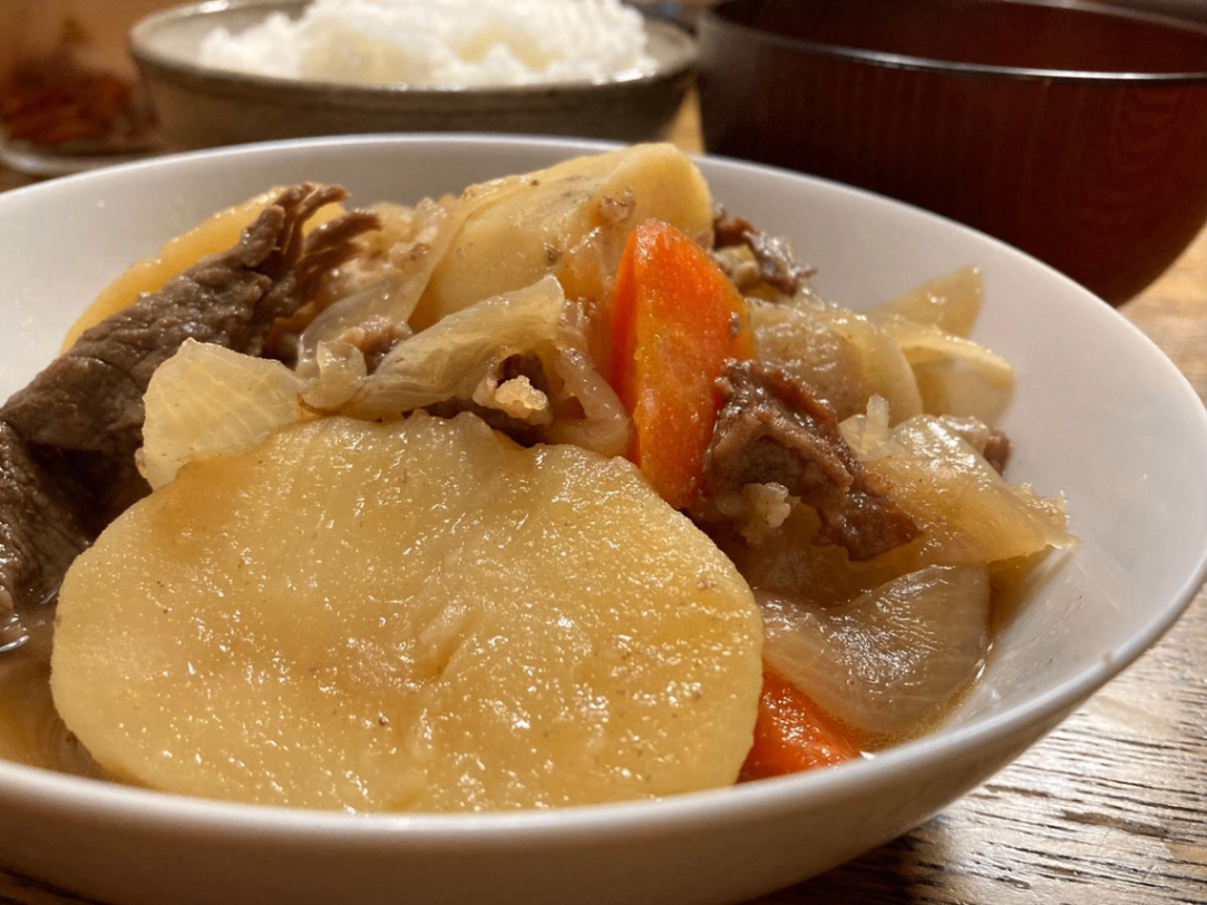 白い器に入った肉じゃが。画面上部(奥)に御飯茶碗、その右に味噌汁茶碗があります。