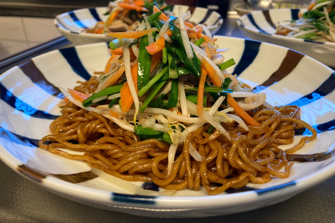 丸いさらに盛り付けた上海焼きそば。焦げ茶色の焼きそば麺の上に、炒めた黒豆もやしと千切りのにんじんとニラと豚肉が盛り付けてあります。