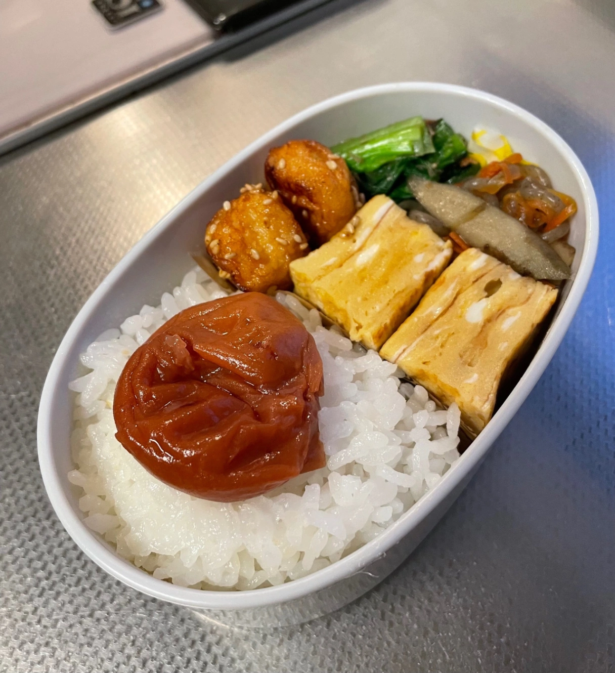 白い楕円形のお弁当箱に詰めたお弁当。メニューは、ごはん、梅干し、卵焼き、玉ねぎ天、ごぼうにんじんこんにゃくのきんぴら、小松菜のお浸し