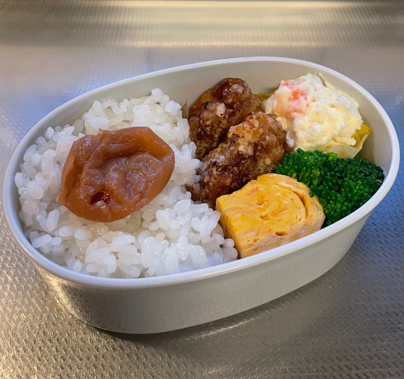 白い楕円形のお弁当箱に詰めたお弁当。メニューは、ごはん、梅干し、カニカマ入り卵焼き、鶏の唐揚げ、茹でブロッコリー、ポテトサラダ