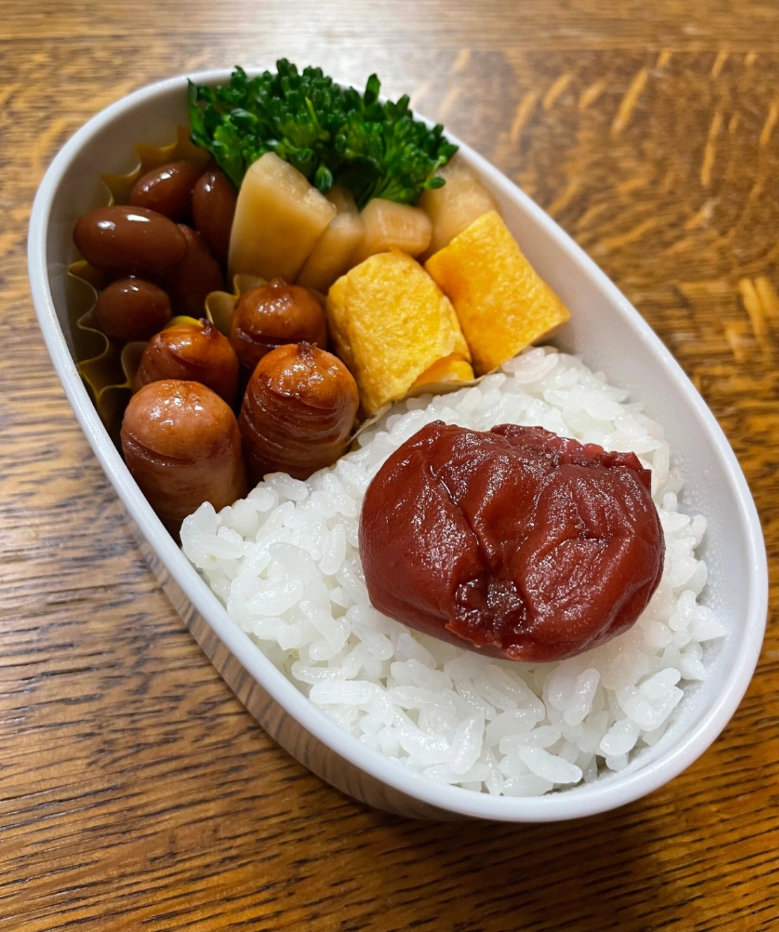 白い楕円形のお弁当箱に詰めたお弁当。献立は、ごはん、梅干し、卵焼き、ウインナー醤油炒め、大根の皮のピリ辛ポン酢漬け、ゆでブロッコリー、金時豆の甘煮