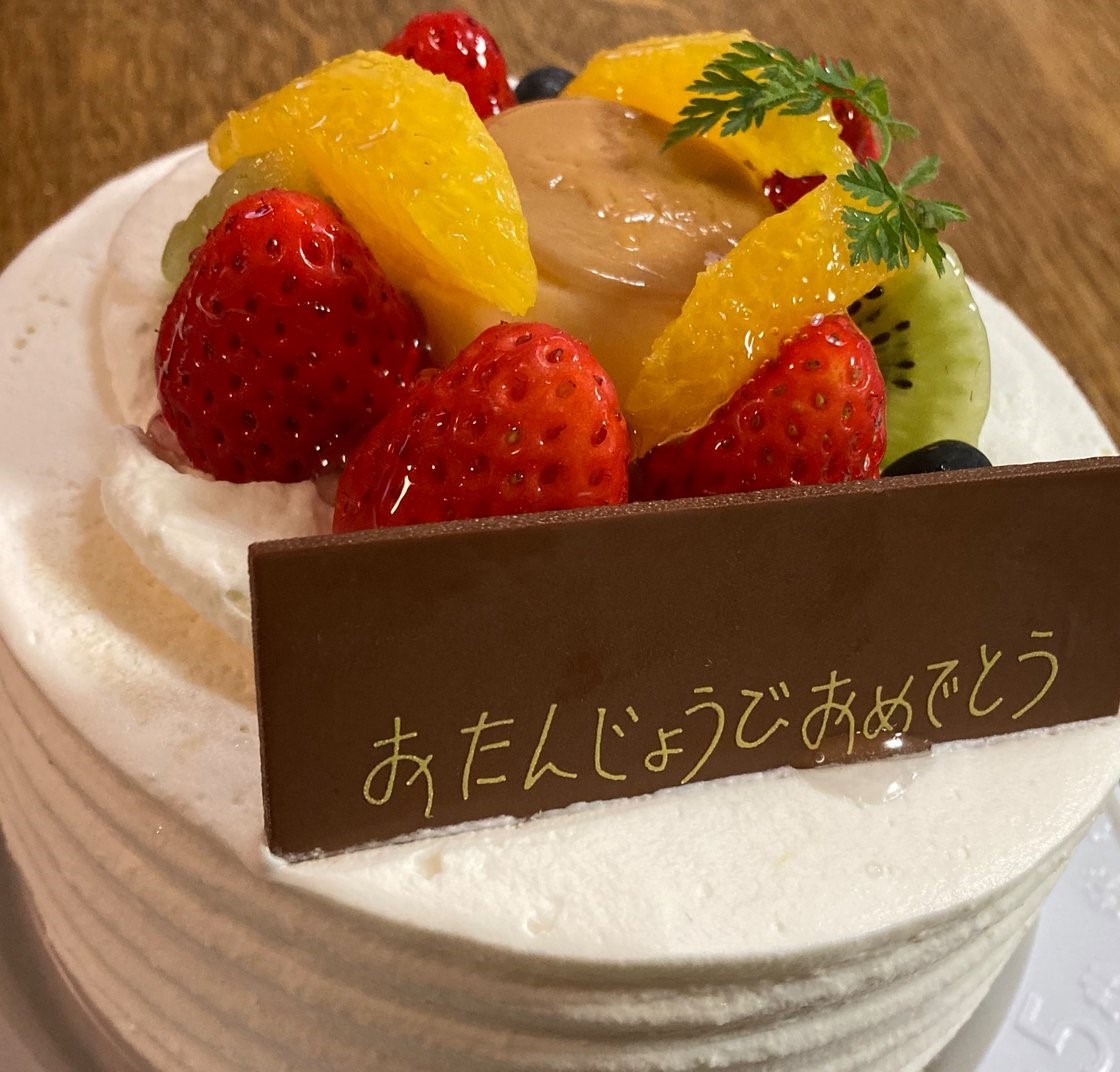 小さなホールのショートケーキ。白い生クリームでコーティングされたケーキの上に小さいプリンとイチゴとミカンとキウイとベリー、それから「おたんじょうびおめでとう」と記されたチョコレートのカードが乗っている。