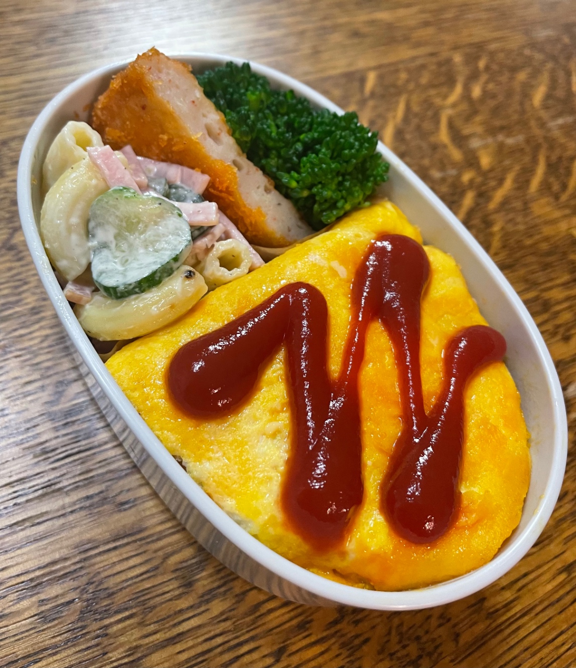 白い楕円形のお弁当箱に詰めたお弁当。献立は、オムライス、茹でブロッコリー、魚ミンチコロッケ、マカロニサラダ