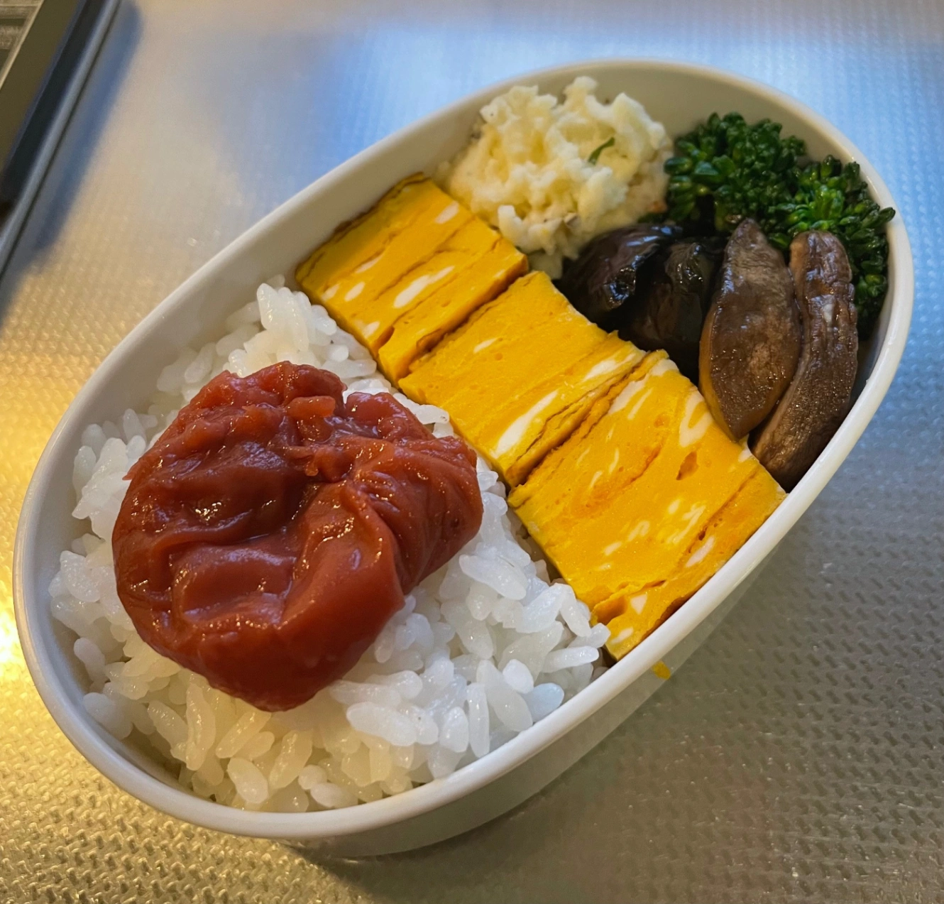 白い楕円形のお弁当箱に詰めたお弁当。メニューは、ごはん、梅干し、卵焼き、ナスの揚げ出し、エリンギとブロッコリーの焼き浸し、大葉とベーコンのポテトサラダ