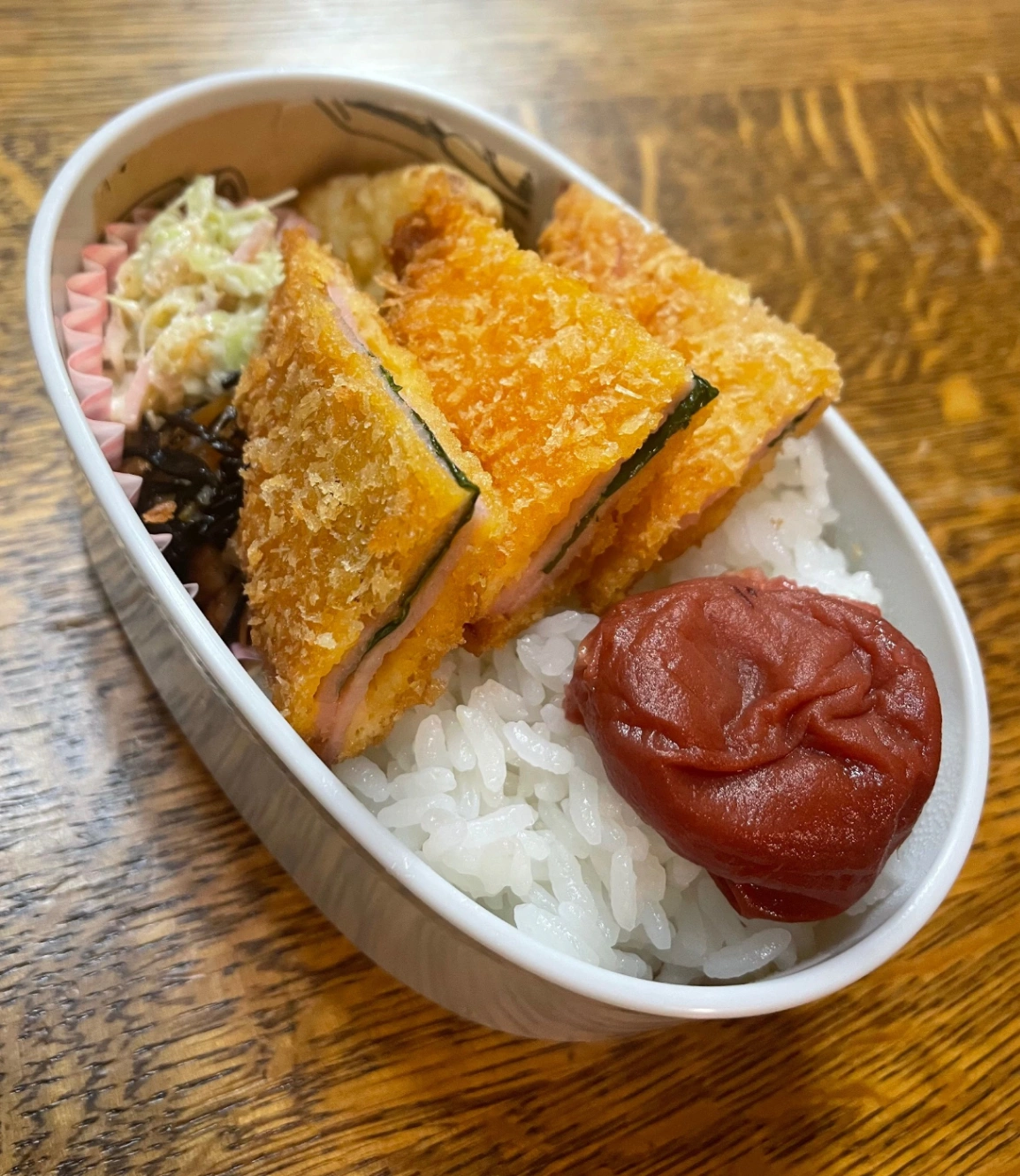 白い楕円形のお弁当に詰めたお弁当。献立は、ごはん、梅干し、大葉入りハムカツ、カリフラワーのフリッター、ひじき煮、キャベツのコールスロー