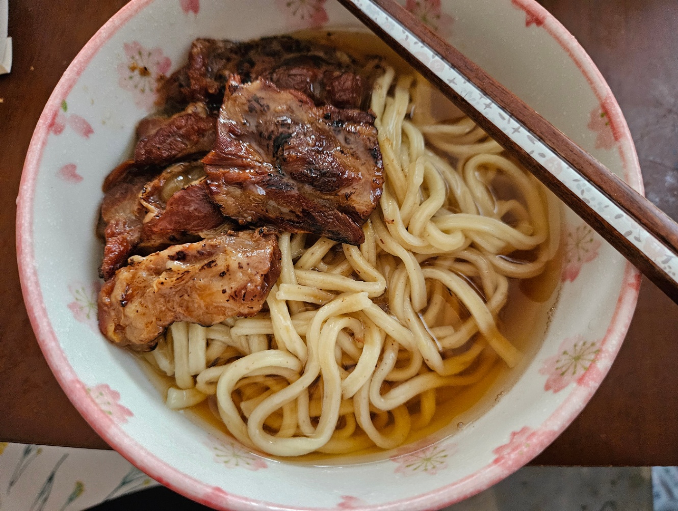 白地にピンクの花がらの丼によそわれたソーキそばの写真。太麺にたくさんの大ぶりのソーキがトッピングされている。お箸が添えられている。