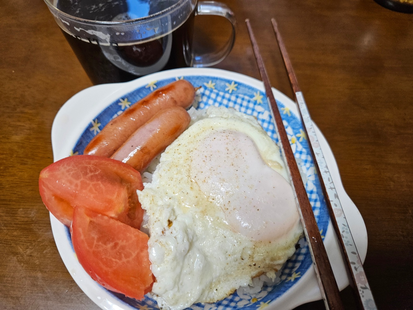 青いチェックで白い持ち手が付いているお皿に盛られた朝ご飯の写真。白ご飯の上に黄身が双子の目玉焼きと、四分の一にカットされたトマト二切れ、ウインナー2本が乗せられている。お箸が添えられていて、透明なマグカップにコーヒーが注がれて置かれている。