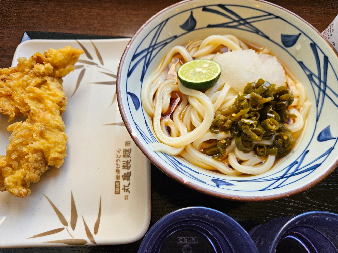 丸亀製麺の青唐おろしうどん(ぶっかけ)の写真。ぶっかけうどんに、大根おろしとすだちの半分、青唐辛子の漬物がトッピングされている。四角いお皿に鶏天が乗っている。それらと水の入ったコップ2つが黒い四角いお盆に乗っている。