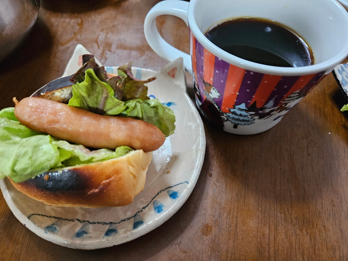 朝ご飯の写真。焼いたロールパンに切れ目を入れてサニーレタスとウインナーを挟んだサンドイッチが猫の形のお皿に乗っている。赤と紫のストライプのムーミンのマグカップにコーヒーが注がれている。