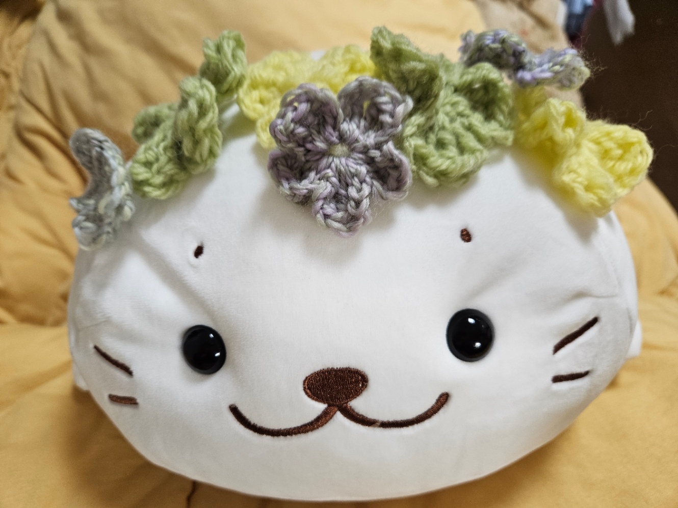 きなこもちしろたんのぬいぐるみの頭の上に黄色や黄緑の毛糸で編んだ花のモチーフがたくさん乗っているところを撮った写真。背景は明るい黄色の掛け布団。