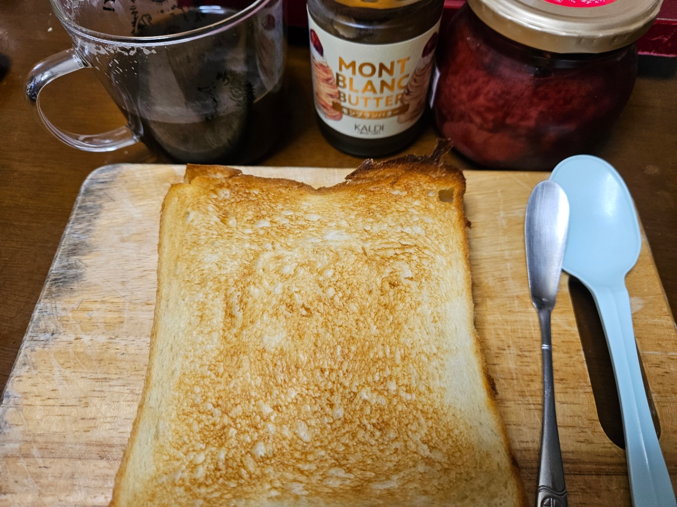 小さい木のまな板の上に乗ったトーストの写真。バターナイフとプラスチックの青いスプーンも乗っている。コーヒーの入った透明なマグカップ、モンブランバターの瓶、苺ジャムの瓶も置いてある。