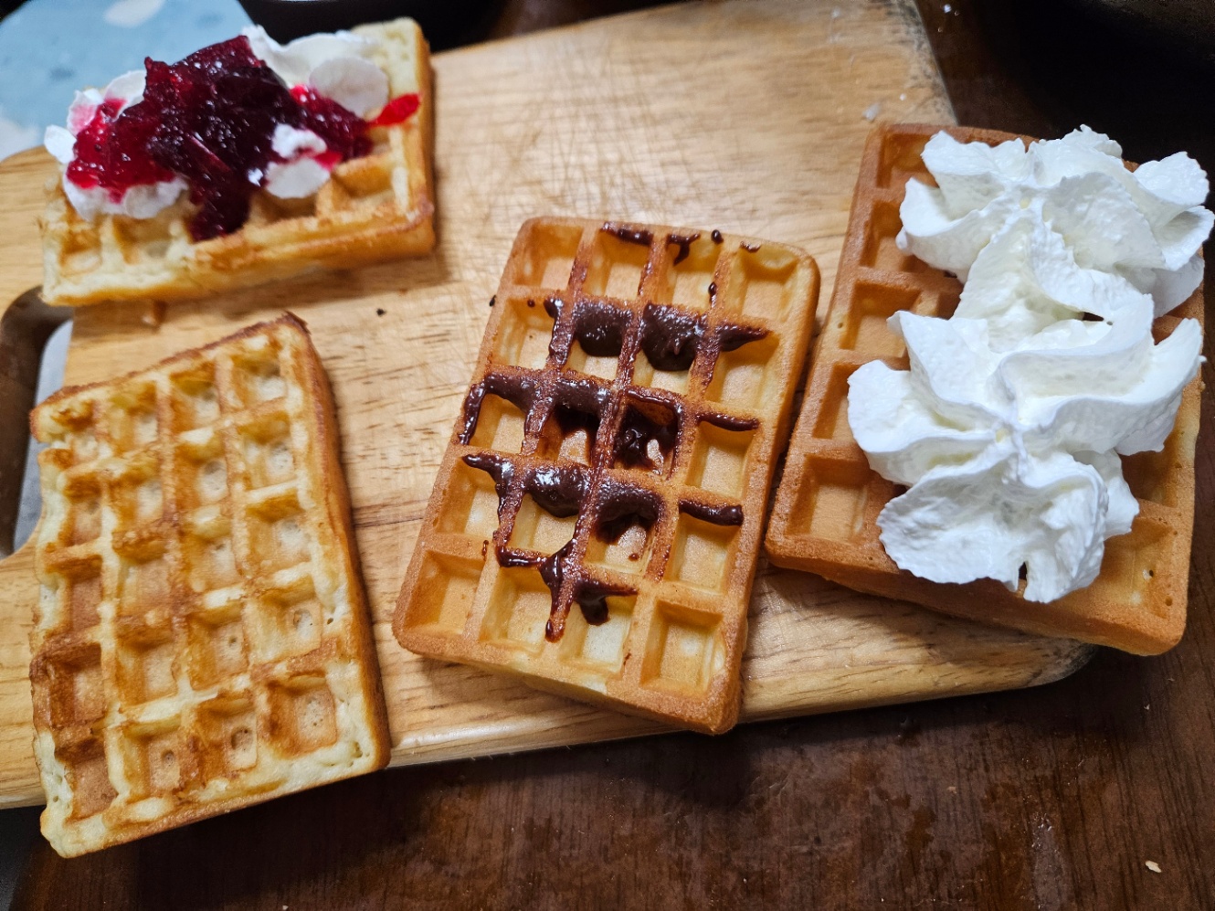 木のまな板の上にワッフルが4個乗っている写真。ワッフルにはそれぞれ生クリームやチョコスプレッドが塗られている。