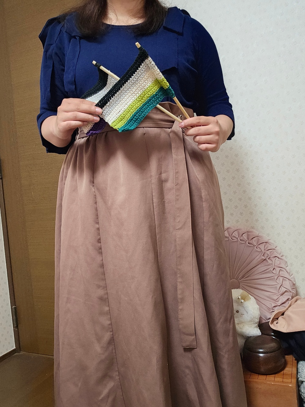 わたしの服装の写真。肩にリボンが付いた紺色の七分袖のニットと、ベルトの付いたココア色のフレアスカートを着ている。手にはアセクシュアルフラッグとアロマンティックフラッグを持っている。