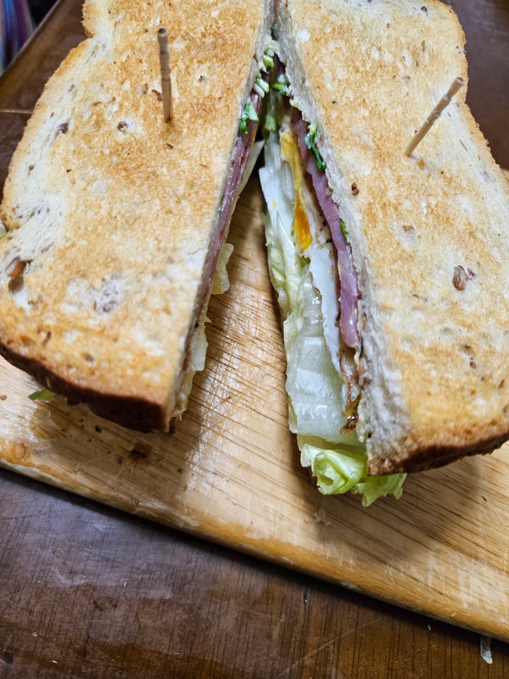 木のまな板に乗ったサンドイッチの写真。焼いたライ麦食パンに、レタス、両面焼き目玉焼き、ベーコン、かいわれ大根が挟んであり、真ん中から切ってある。それぞれ爪楊枝が刺さっていて、断面が見えている。