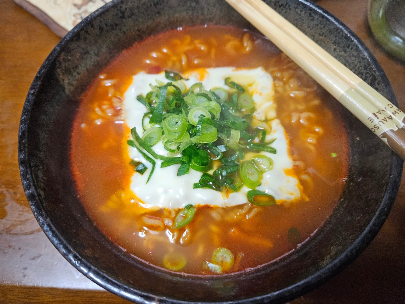 黒い丼によそわれた辛ラーメンの写真。スライスチーズと刻みネギがトッピングされていて、お箸が添えられている。