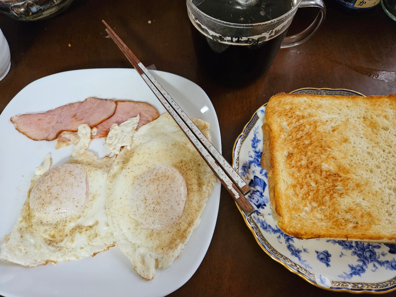 朝ご飯の写真。白い角の丸い四角いお皿に目玉焼き二切れとハム2枚が乗っていて、お箸が添えられている。白地に青い花がらのお皿にトーストが一枚乗っていて、透明なマグカップにコーヒーが注がれて置いてある。