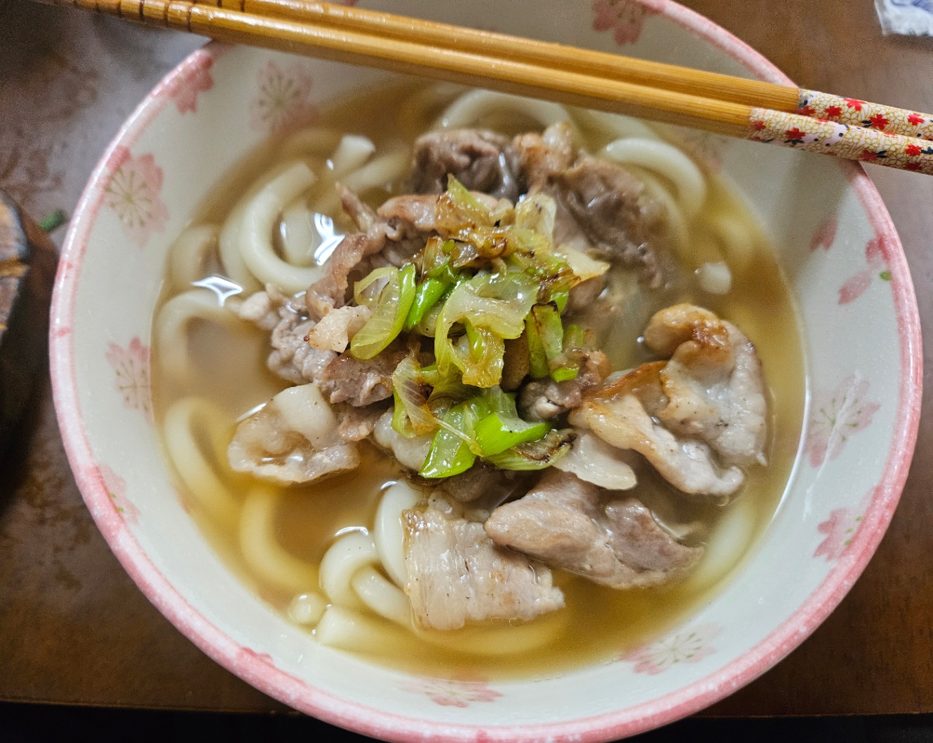 白地にピンクの花がらの丼にうどんがよそってあるところを撮った写真。炒めた豚肉と長ネギがトッピングされていて、お箸が添えられている。