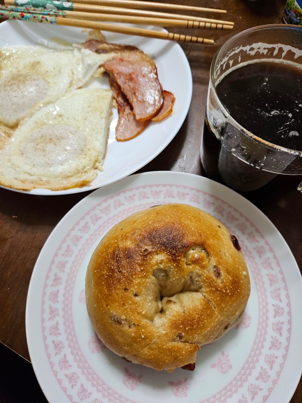 朝ご飯の写真。白い丸いお皿に目玉焼き二切れとハム4枚が乗っていて、手前の白地にピンクの花がらのお皿にはクランベリーとマカダミアナッツのベーグルが焼かれておいてある。透明なカップにコーヒーが注がれて置いてある。