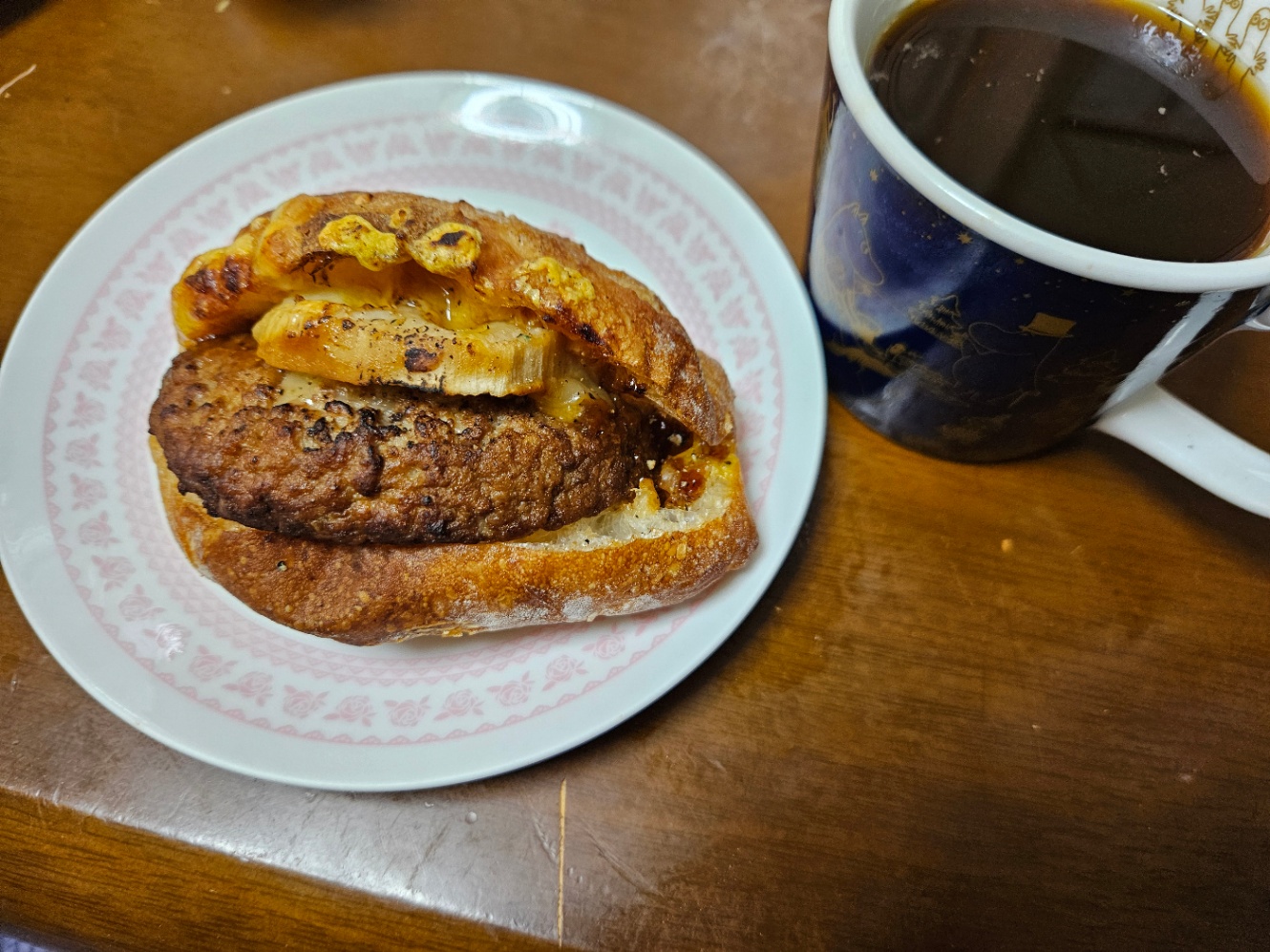 白地にピンクの花がらのお皿に乗った惣菜パンの写真。ハンバーグとれんこんが挟んであるリュスティック。紺色のムーミン柄のマグカップにコーヒーが注いでおいてある。