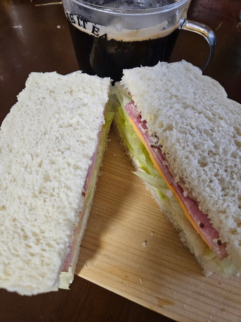 木のまな板の上に乗っているサンドイッチの写真。耳の白い食パンで、レタス、ハム、スライスチェダーチーズが挟んである。透明なマグカップにコーヒーが注いでおいてある。