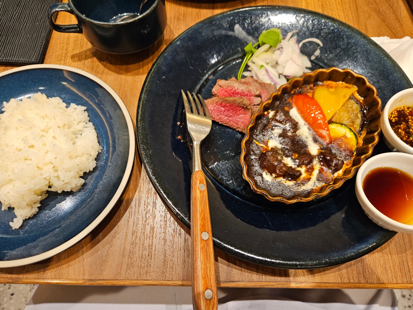 お昼ごはんの写真。左手にお皿に盛り付けられた白ご飯、右手にビーフシチューの器と三枚のローストビーフ、ソースの器2つが乗った大きなお皿がおいてある。奥にはスープのカップもある。