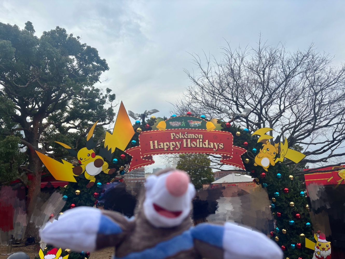 横浜赤レンガ倉庫のポケモンブースにあるアーチの写真。メガシンカしたライチュウのパネルが左右にあり「Pokémon happy holidays」と書かれたリボン型の看板が中央に飾られています。Pokémon fitのモグリューのぬいぐるみが手前に写っています。